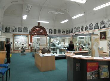 Image shows the inside of Hereford Museum with glass display cases around the outside of the large room, and table top displays in the centre of the room.