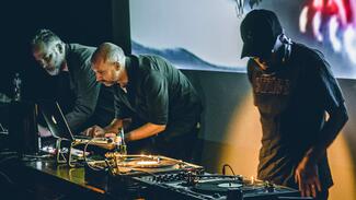 Three men using decks and a laptop with a screen behind them