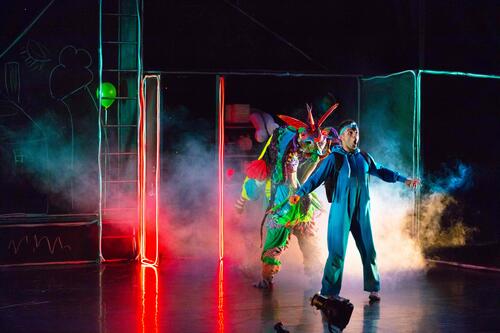 Actors in multi-coloured costumes on a dark stage lit by red light